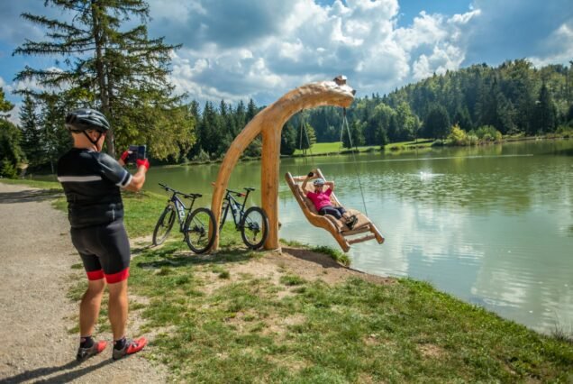 Biker couple enjoying and taking photos at a wooden swing over Lake Bloke at Nova Vas in Slovenia