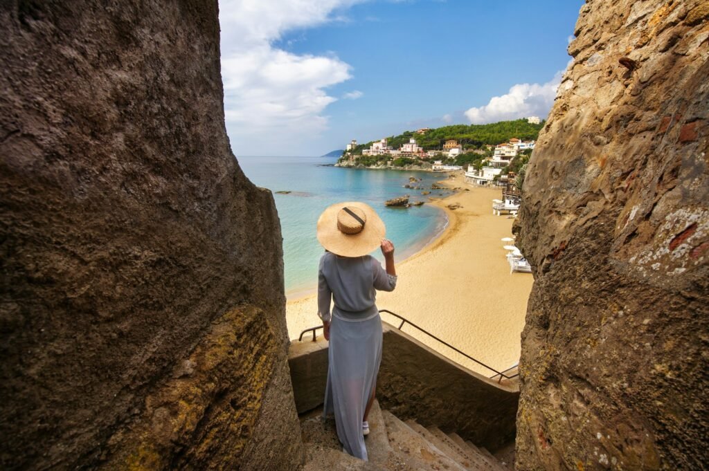 A girl in a hat stands on a mountainside overlooking the sea and the beach in Italy