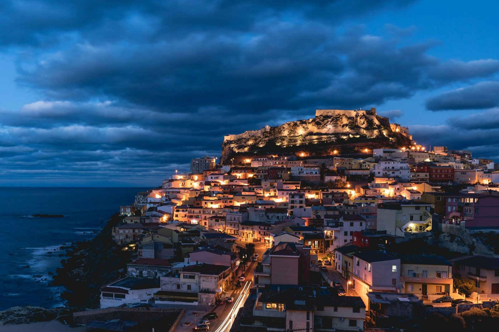 Aerial cityscape of Castelsardo surrounded by buildings and lights near water in evening